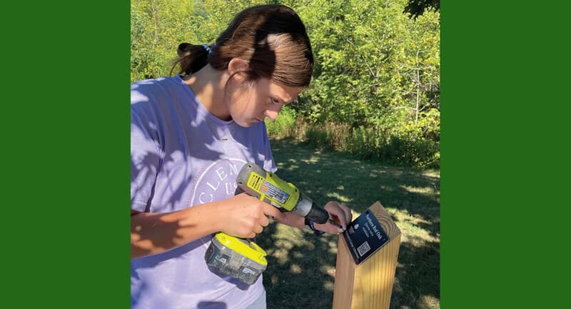 Photo of a student screwing a placard onto a wooden post.