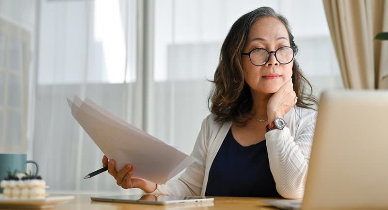 Photo of a woman looking at a laptop and holding a pen and paper